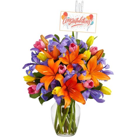 A colorful bouquet in a glass vase featuring orange lilies, purple irises, and yellow and pink tulips. A card with "Congratulations!" is perched on a stick above the bouquet.