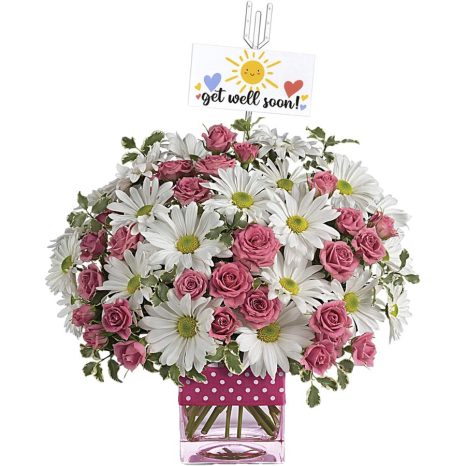 A floral arrangement featuring white daisies and pink roses in a square vase with a pink polka dot ribbon. A card with "get well soon!" and a smiling sun, hearts, and flowers is attached at the top.