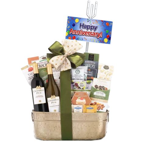 A metal basket filled with two bottles of wine, snacks, pasta, and sauces, decorated with a green ribbon and bow. A "Happy Anniversary" sign is attached to the basket with a festive design.