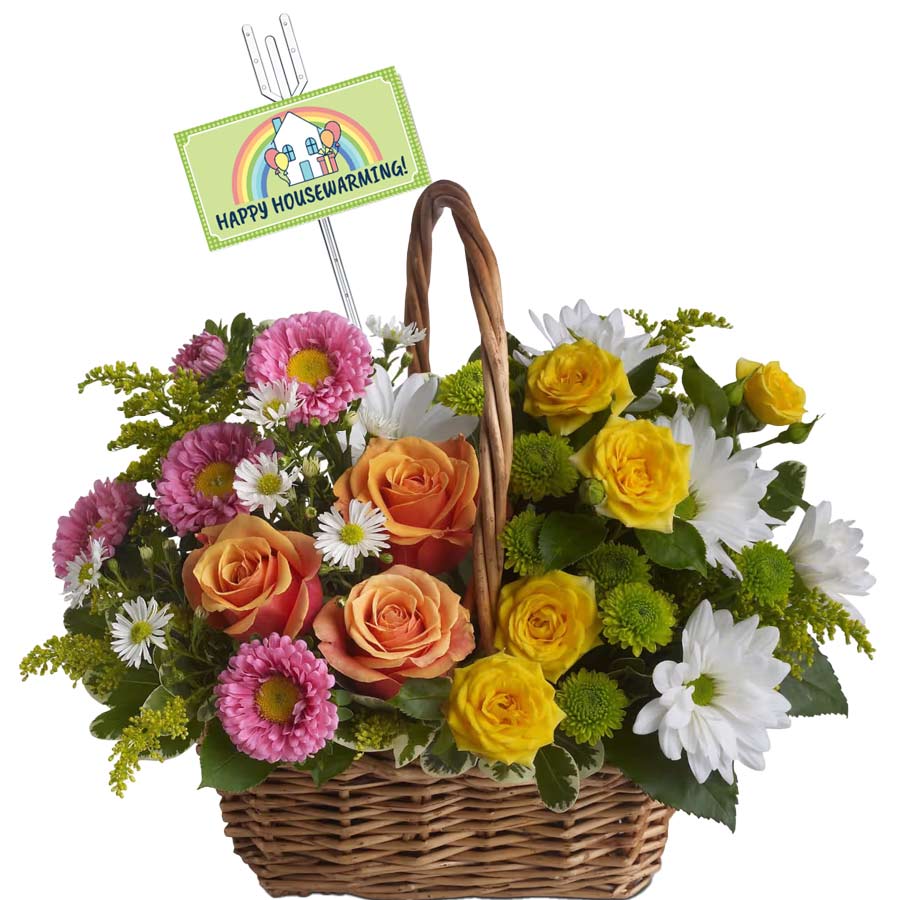 A wicker basket filled with a colorful arrangement of pink, yellow, and peach flowers, including roses and daisies. A sign sticks out of the bouquet with the text "Happy Housewarming!" and a rainbow graphic.