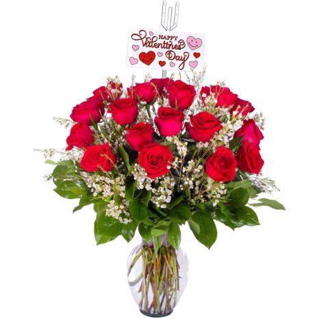 A bouquet of red roses in a glass vase, adorned with small white flowers and green leaves. A "Happy Valentine's Day" card with hearts is clipped to the arrangement.